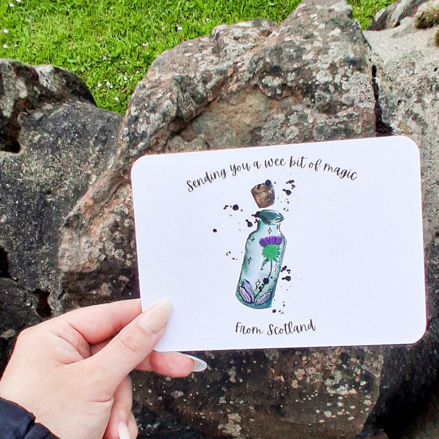Hand holding a card with a bottle illustration and text, set against a natural rock and grass background.
