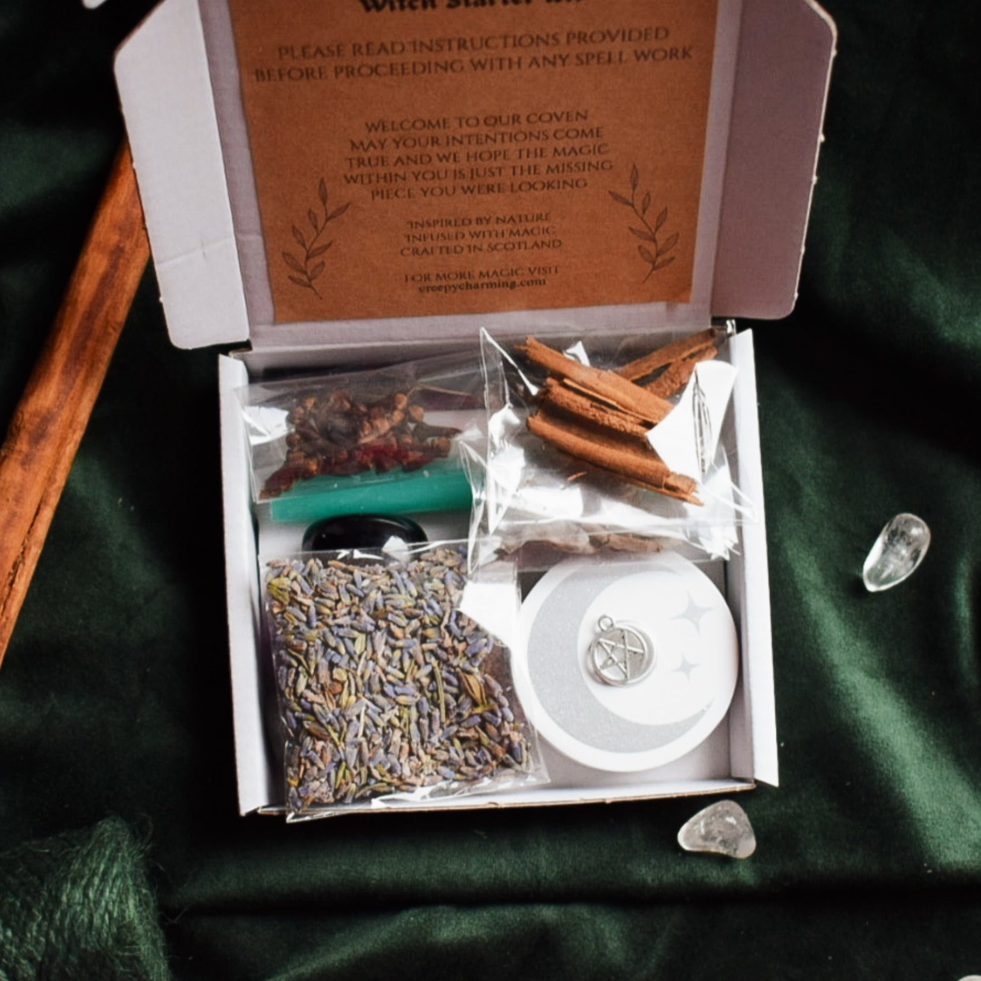 Box of spell components including lavender, cinnamon sticks, and a green candle on a dark green fabric background.